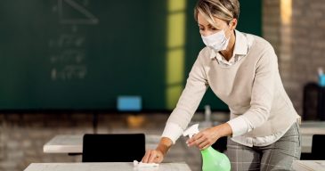 Teacher with face mask preparing classroom for kids and cleaning tables with disinfectant.